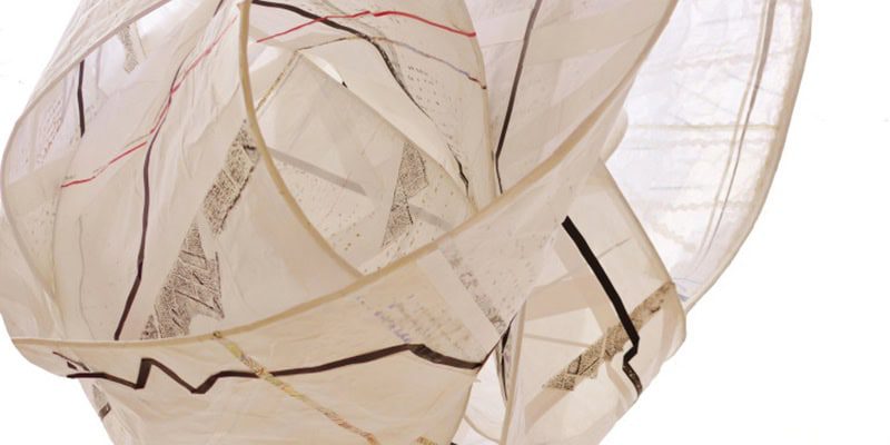 A macro detail of a sculptural paper installation. The work is composed of overlapping, translucent white paper bands with thin black and red lines. Hand-drawn text and geometric markings are visible through the layers, creating a complex, nested form.