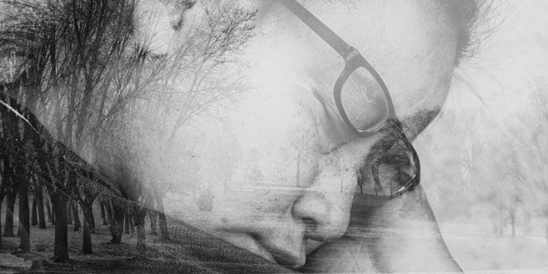 A monochromatic double-exposure photograph. The soft-focus profile of an individual wearing glasses is overlaid with the fine, skeletal branches of a winter forest. The textures of the skin and the trees merge, creating an ethereal, contemplative mood.