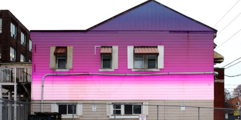A digital artwork of a two-storey house with a gabled roof. The upper half of the building is saturated in a deep violet and magenta gradient, which transitions into a bright, glowing horizontal band of neon pink across the middle, fading into soft peach near the ground.