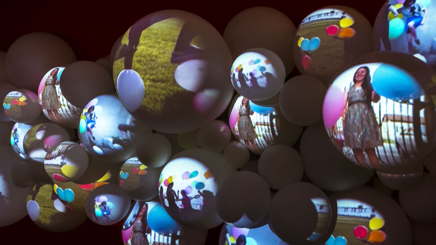 A close-up of a multimedia installation featuring several white, spherical balloons of various sizes. Video imagery is projected onto the spheres, showing individuals holding colourful balloons and silhouettes.