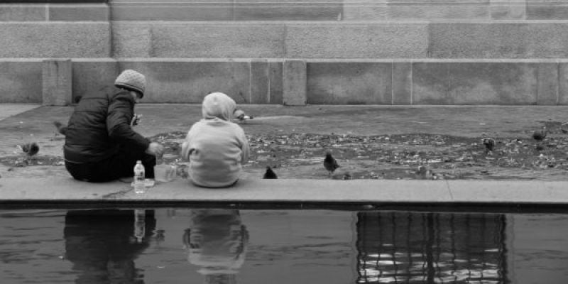 A monochromatic landscape photograph from "The Third Place." Two individuals sit on a low concrete ledge by a reflecting pool, feeding a flock of pigeons. Their reflections are visible in the still water in the foreground.