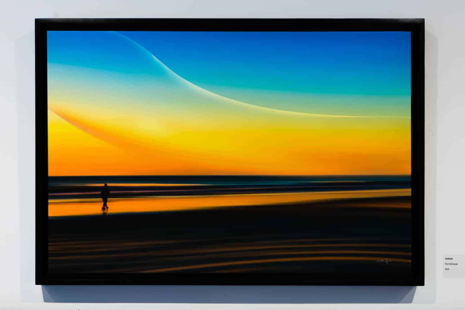 A framed horizontal photograph titled "Solitude" by Rick McKenzie. A vibrant sunset scene simplified by long exposure into bands of brilliant orange, yellow, and cyan. A single dark silhouette of a person walks along the shore, reflected in the wet sand.