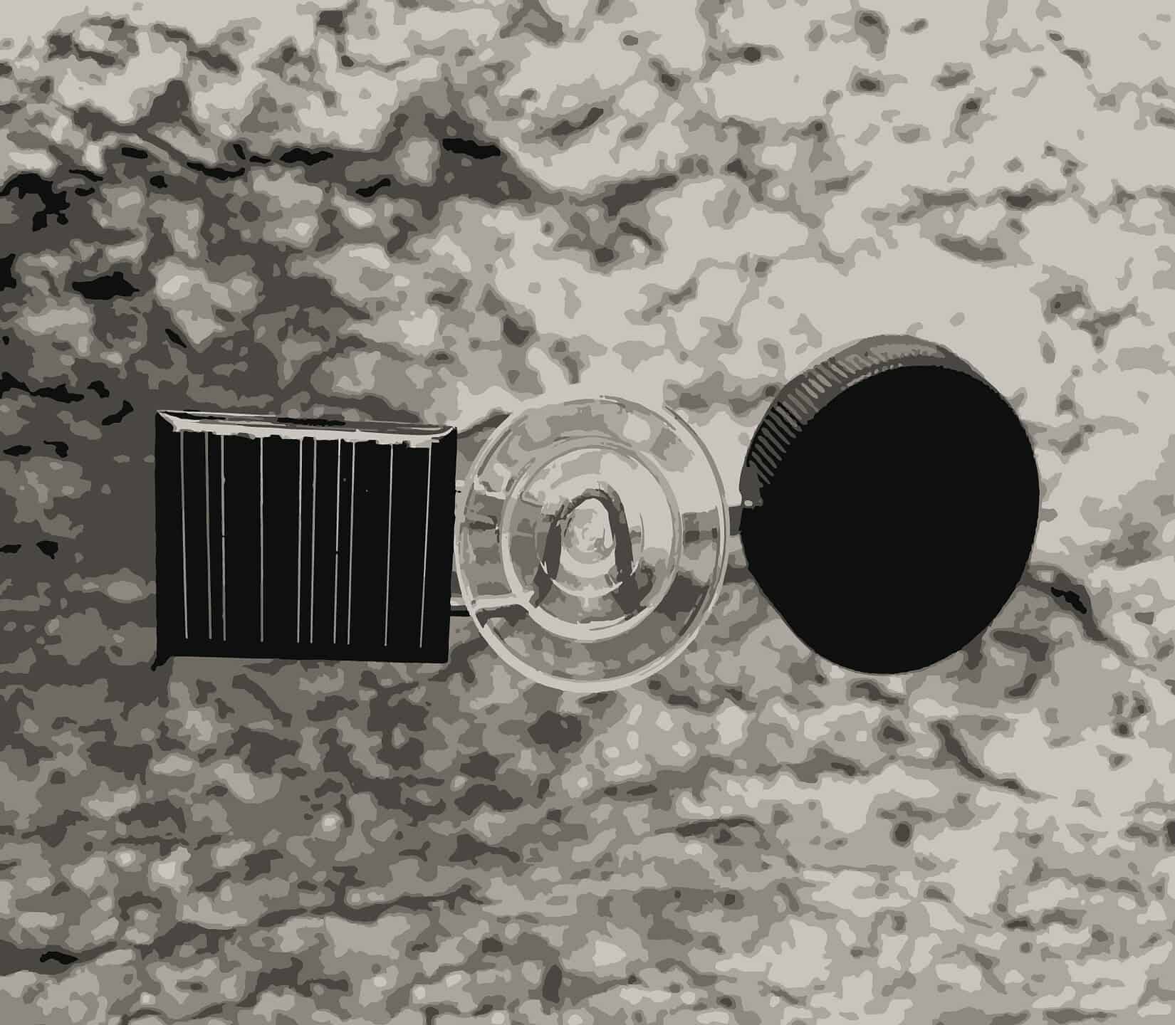 A stylized, high-contrast graphic of the "Reality Gap" installation components. It shows a small solar panel, a circular suction cup, and a black round cap arranged horizontally against a textured, grey-toned background.