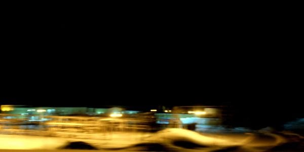 A panoramic long-exposure photograph taken at night. Distant buildings and lights are transformed into glowing horizontal streaks of yellow and white across a dark, snow-covered landscape under a vast black sky.