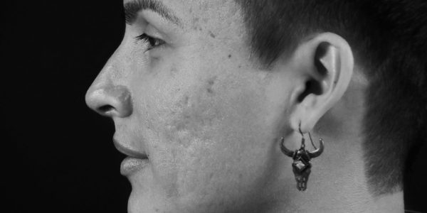 A black-and-white profile portrait of a person with short hair. The lighting emphasizes the texture of their skin and a unique earring shaped like a detailed animal skull with horns.