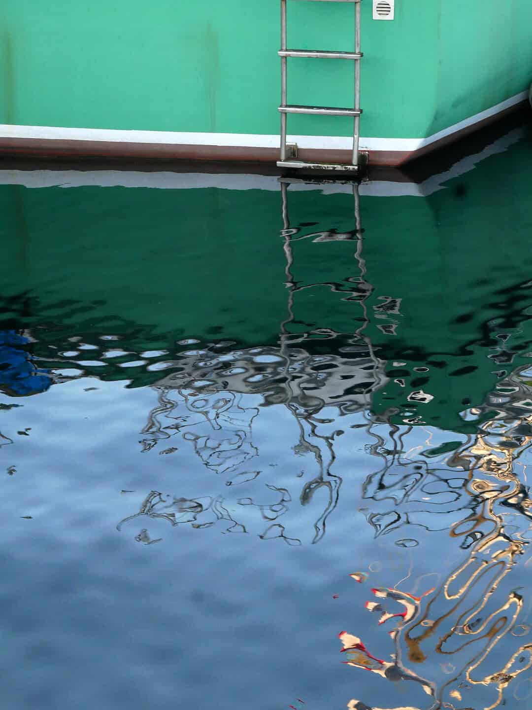 A vertical photograph capturing the reflection of a green boat and a silver metal ladder in rippling dark water. The abstract, distorted reflections create a rhythmic pattern of blue, green, and white.