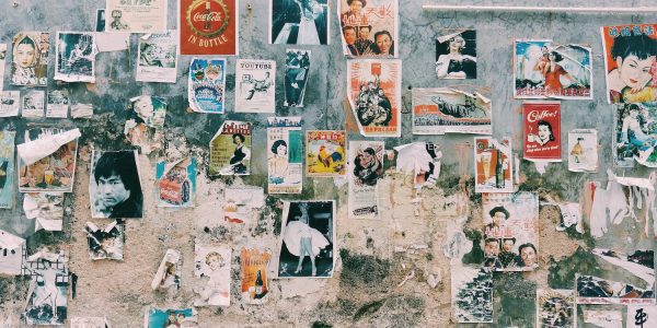 A wide photographic mural of a weathered urban wall covered in a dense collage of vintage posters, advertisements, and torn paper scraps. Images of iconic figures like Bruce Lee and Marilyn Monroe are visible among various typographic styles.