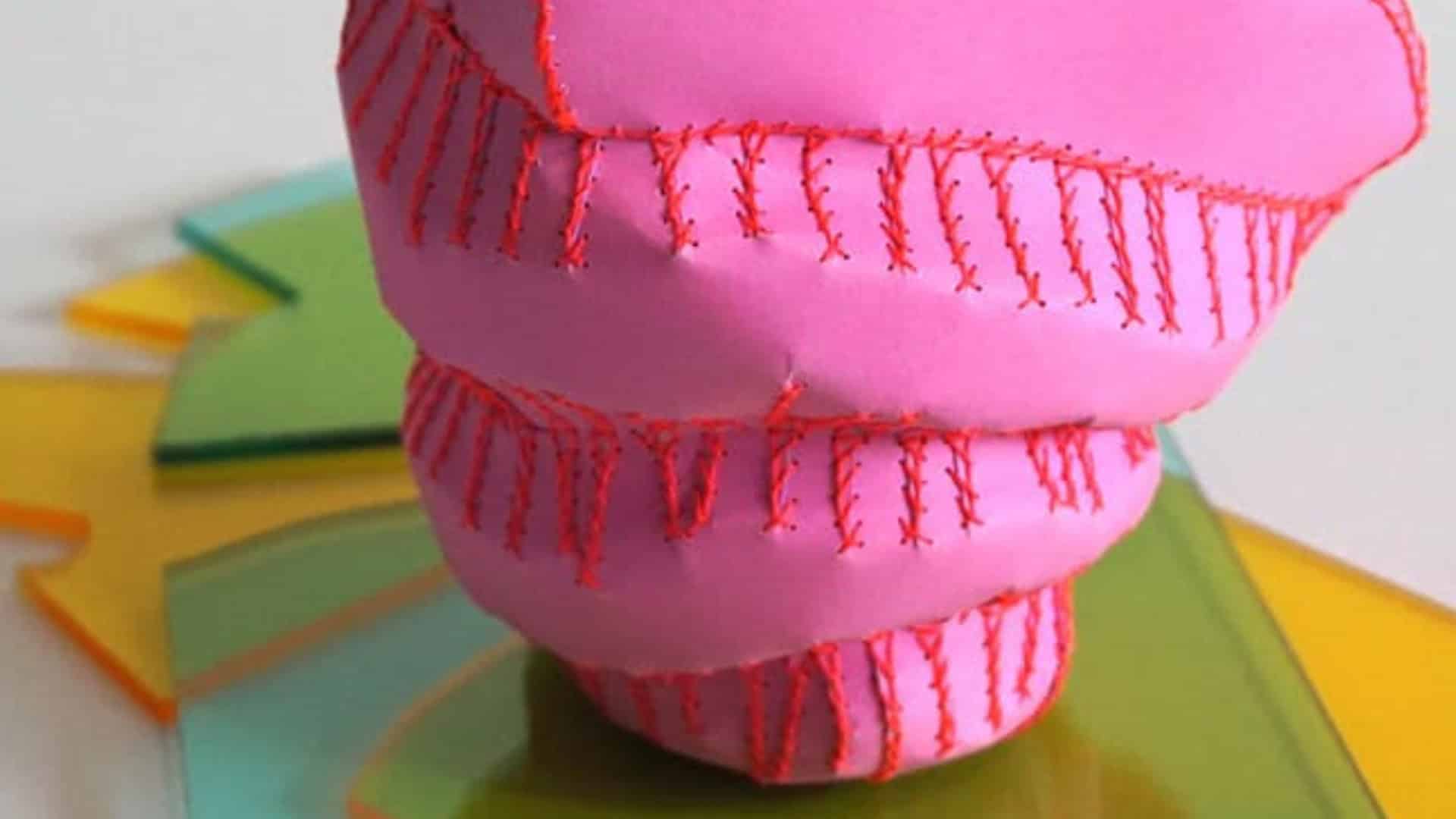 A close-up of a soft sculpture made of bright pink, matte material. The form is bulbous and tapered, reminiscent of a shell or organic pod, with prominent, thick red stitching creating ridges across its surface. It sits atop colourful, translucent acrylic sheets.