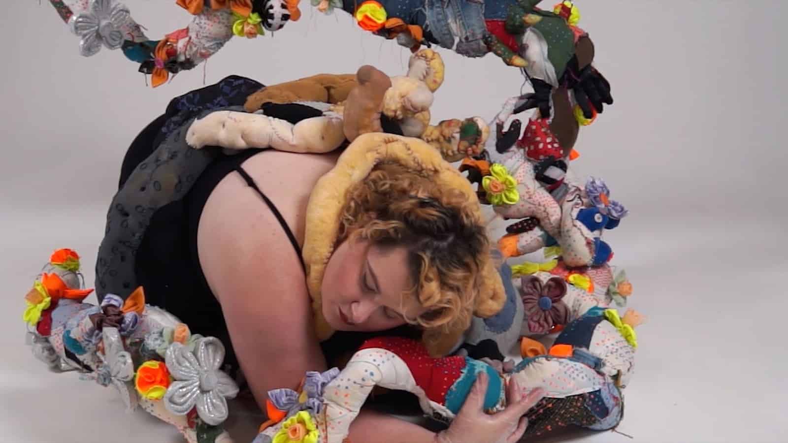 Person with short curly hair crouched on the floor, partially encircled by a large soft sculptural form made of stuffed fabric segments decorated with bright felt flowers and small objects, against a plain white background