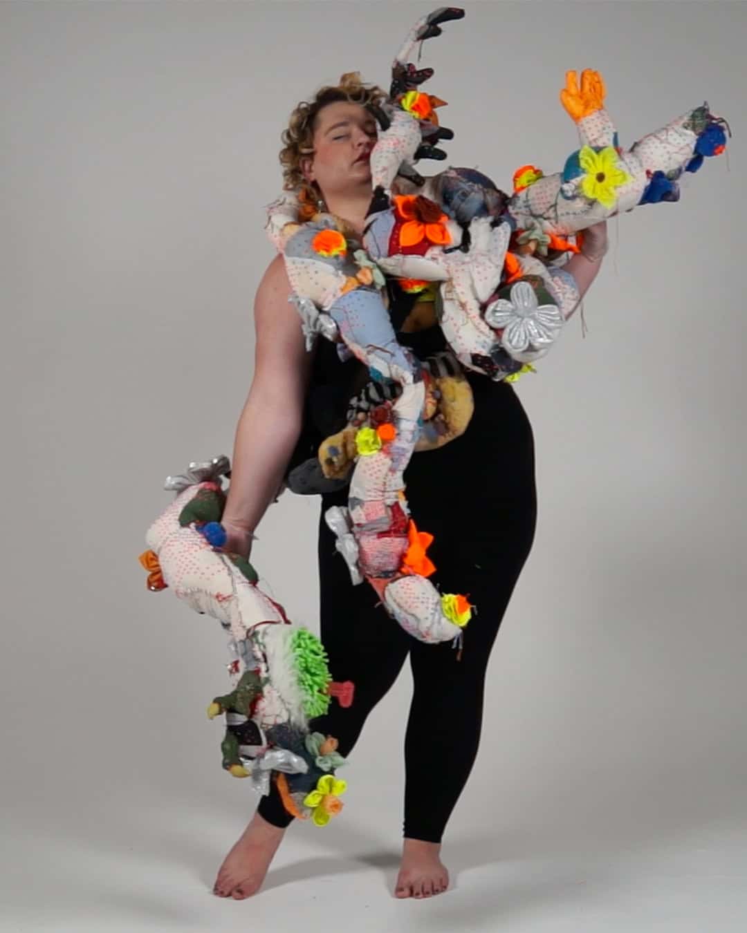 Person standing barefoot in a black outfit, draped with a long, multi-limbed soft sculpture made of patterned fabric and bright appliqué flowers, extending from shoulders and arms against a white backdrop