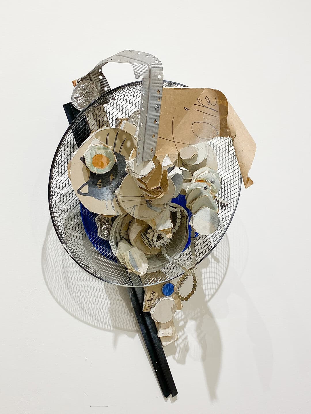 Circular wire mesh assemblage filled with layered cardboard, painted fragments, and small objects, with a curved perforated metal strip arching over the top