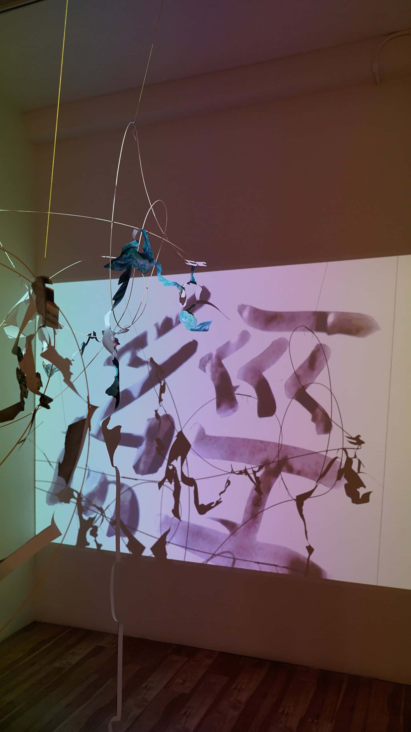 Suspended installation casting layered shadows over projected abstract calligraphic forms on a gallery wall