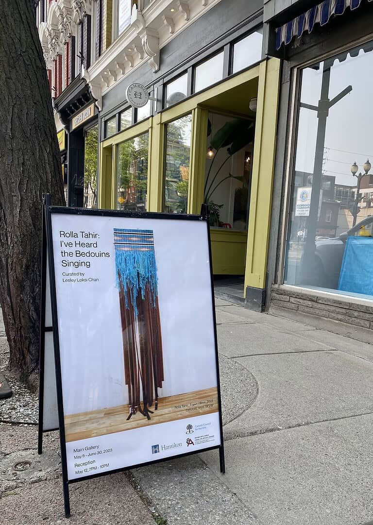 Sidewalk sandwich board sign outside a gallery displaying a poster for “I’ve Heard the Bedouins Singing” by Rolla Tahir