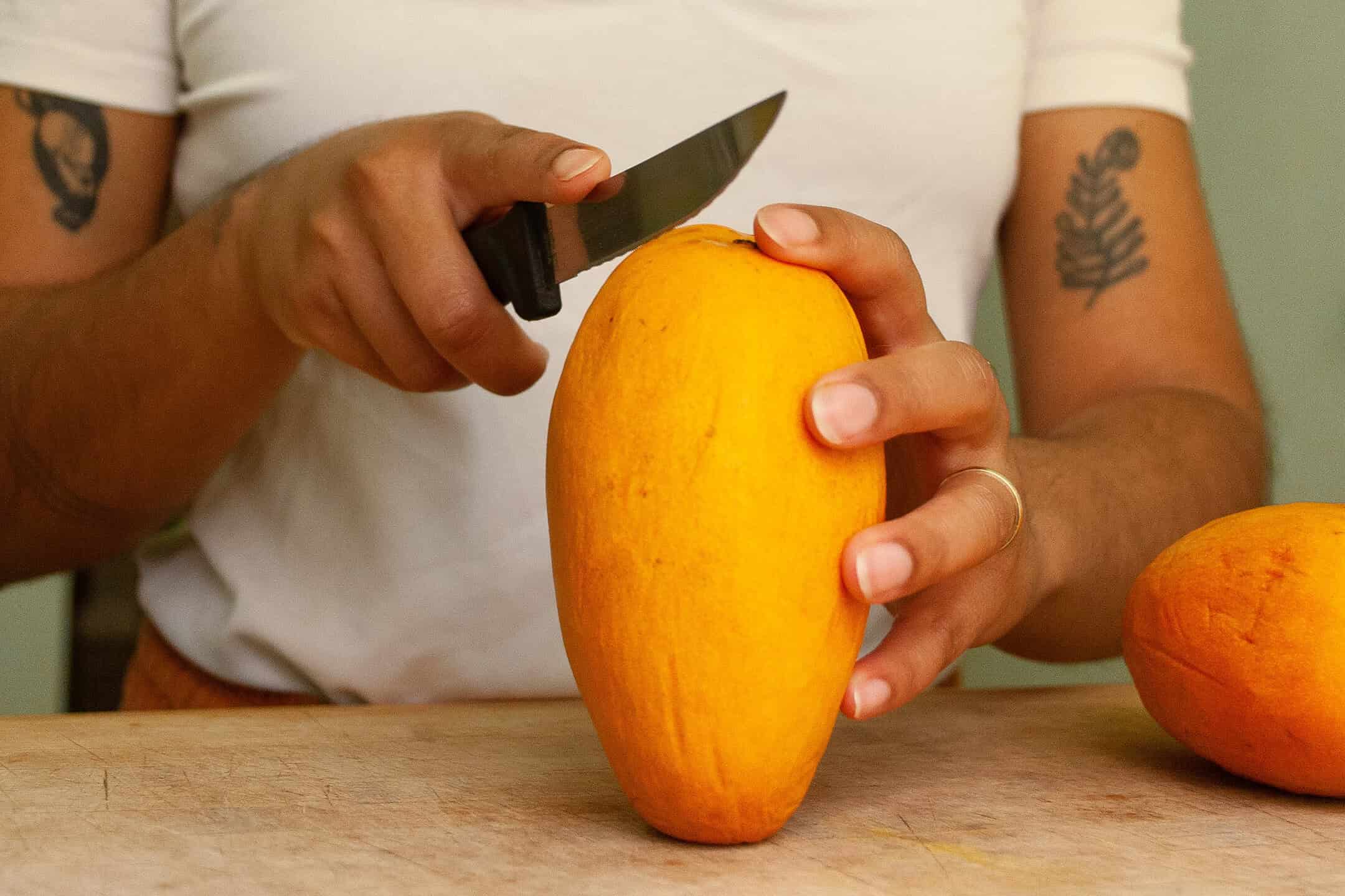 Close-up photograph of hands holding a mango while slicing it with a knife on a cutting board