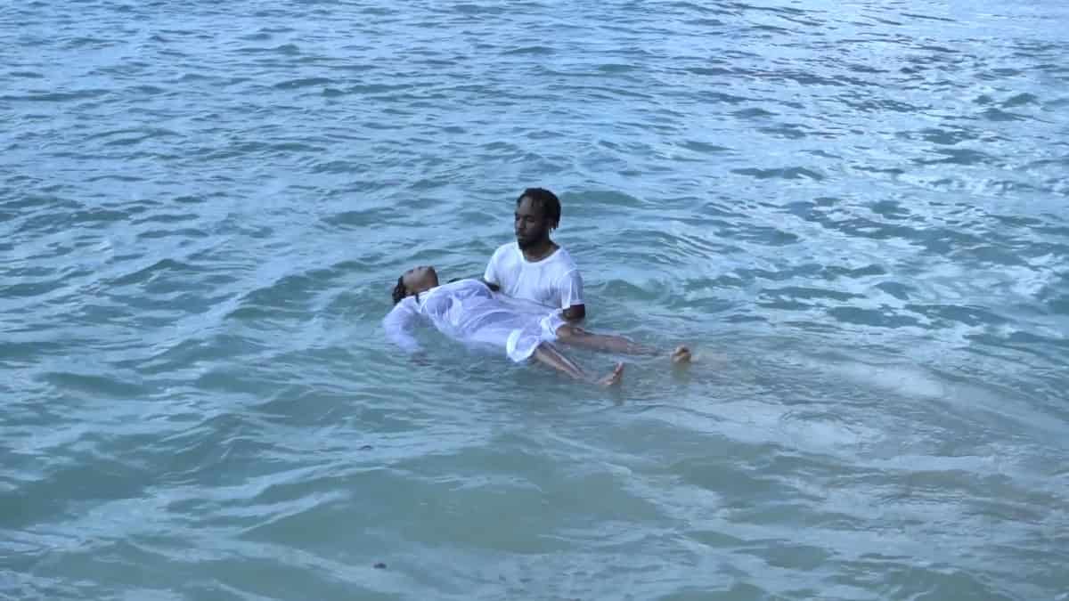 Two people in shallow ocean water, one holding another who is floating on their back, both dressed in light clothing