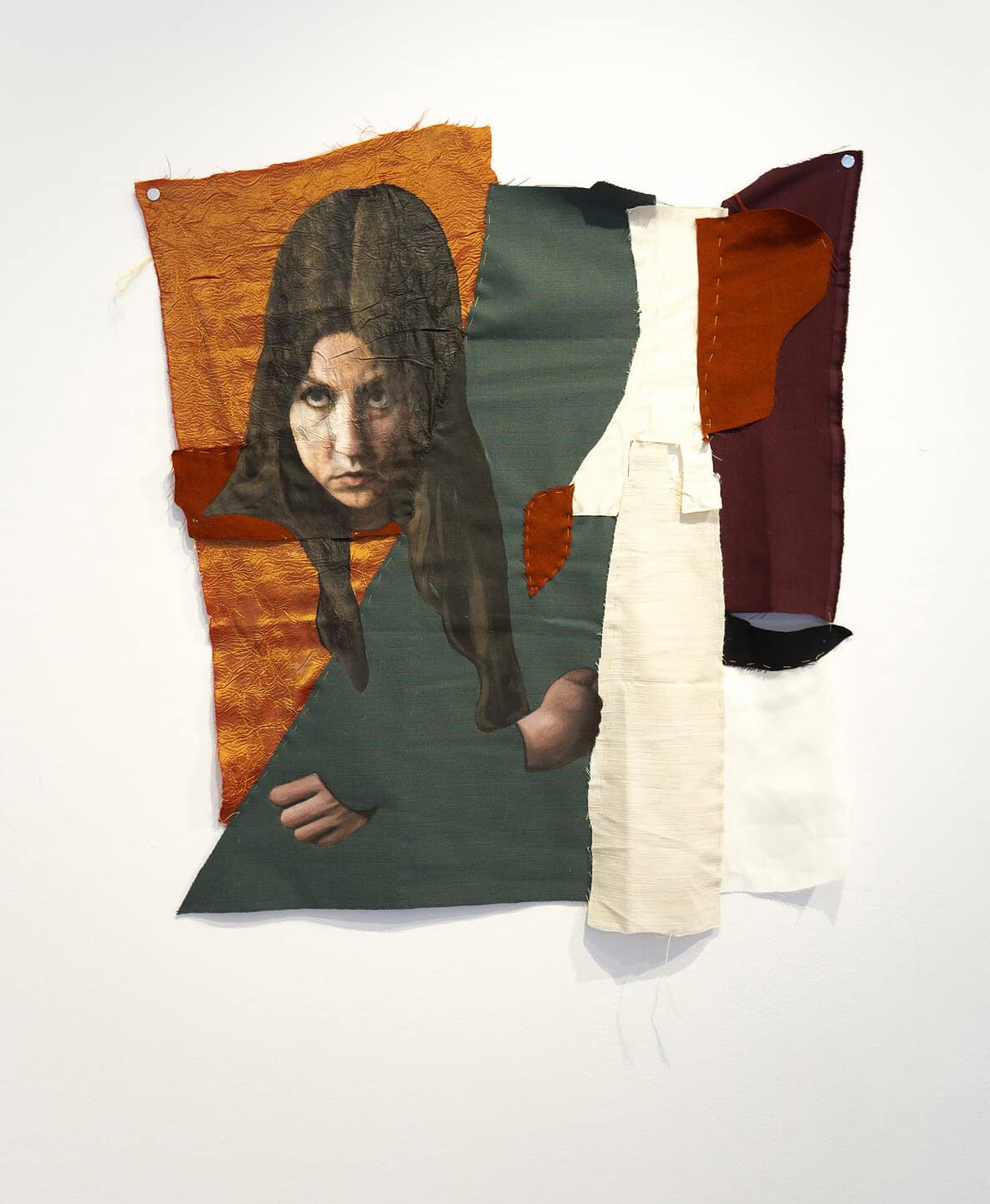 Textile artwork with layered orange, green, and cream fabrics framing a painted face looking forward
