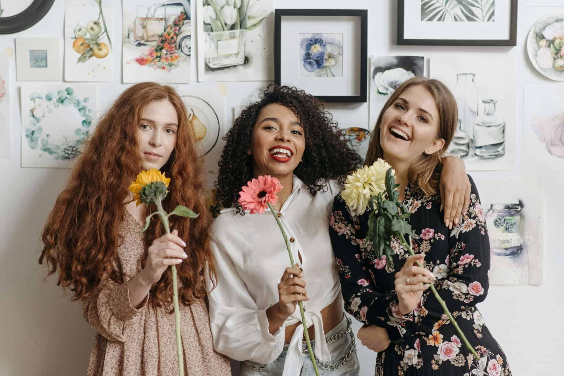 Three stylish young people smiling and holding cut flowers stand before a wall adorned with artistic prints, reflecting the vibrant spirit of Canadian Artist-Run Centres.