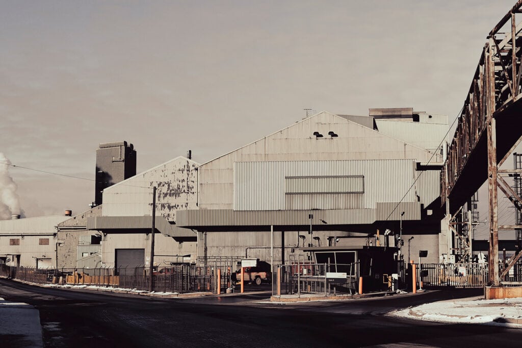 A photograph of large industrial factory in Hamilton with metal siding and smokestacks, surrounded by a fence. There is some snow on the ground and a clear sky in the background.