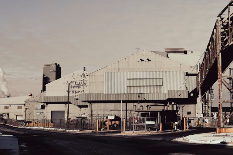 A photograph of large industrial factory in Hamilton with metal siding and smokestacks, surrounded by a fence. There is some snow on the ground and a clear sky in the background.