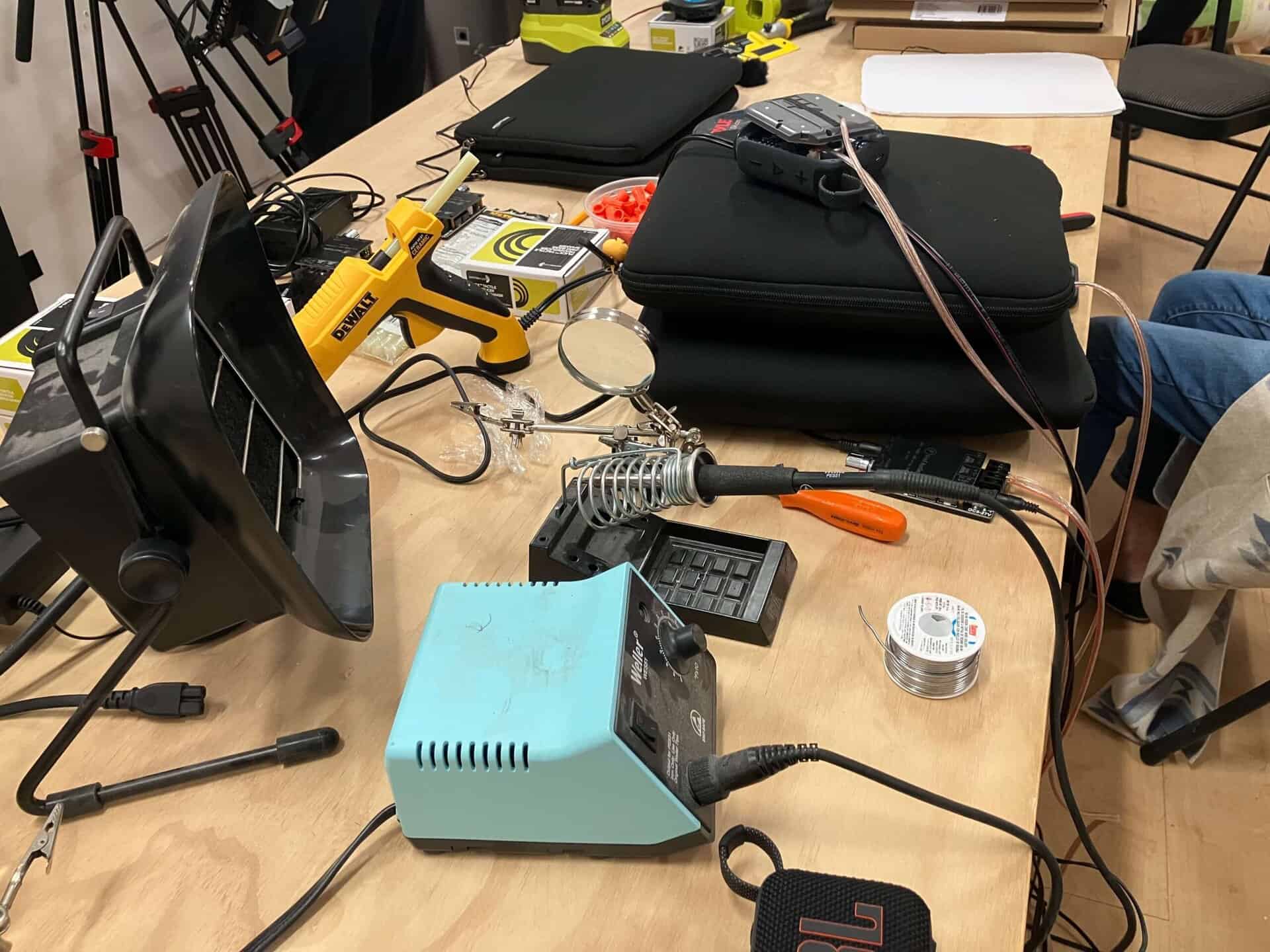 A workshop table cluttered with electronics tools, including a soldering iron, solder wire, haptic technologies components, a glue gun, wires, black cases, pliers, and a magnifying glass on a wooden surface.