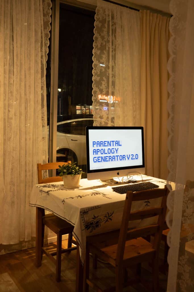A computer on a small dining table displays the text PARENTAL APOLOGY GENERATOR V 2.0. Surrounded by a plant and lace curtains the table sits by a window, with a car visible outside at night.