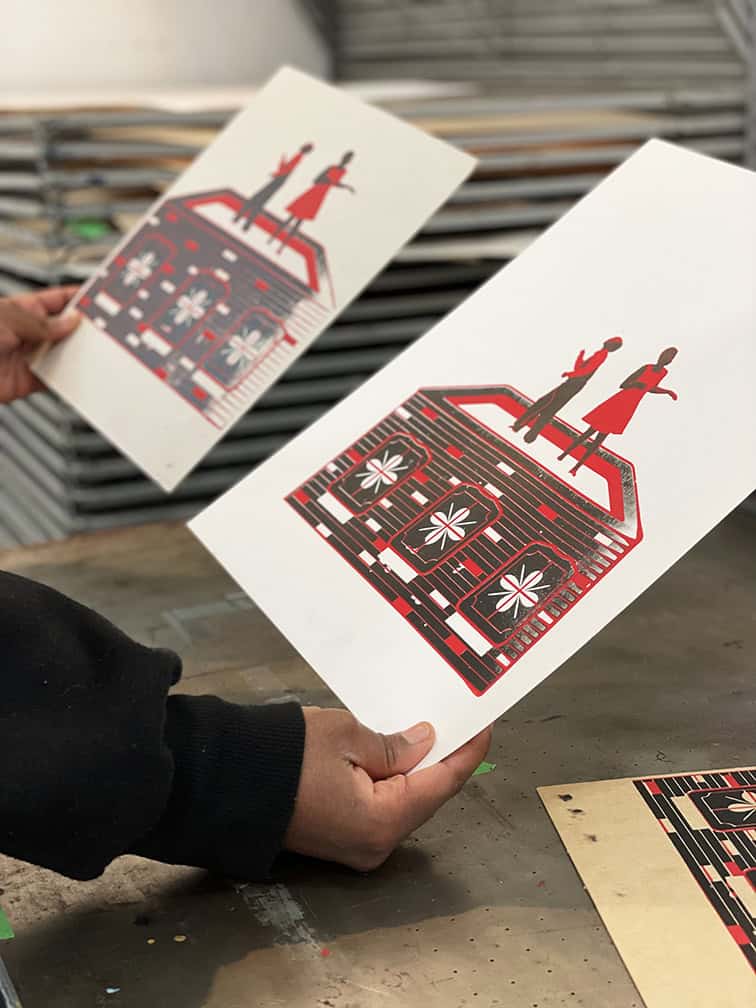 A person holds two matching screen prints showing stylized red and black images of people standing on a house with decorative patterns.