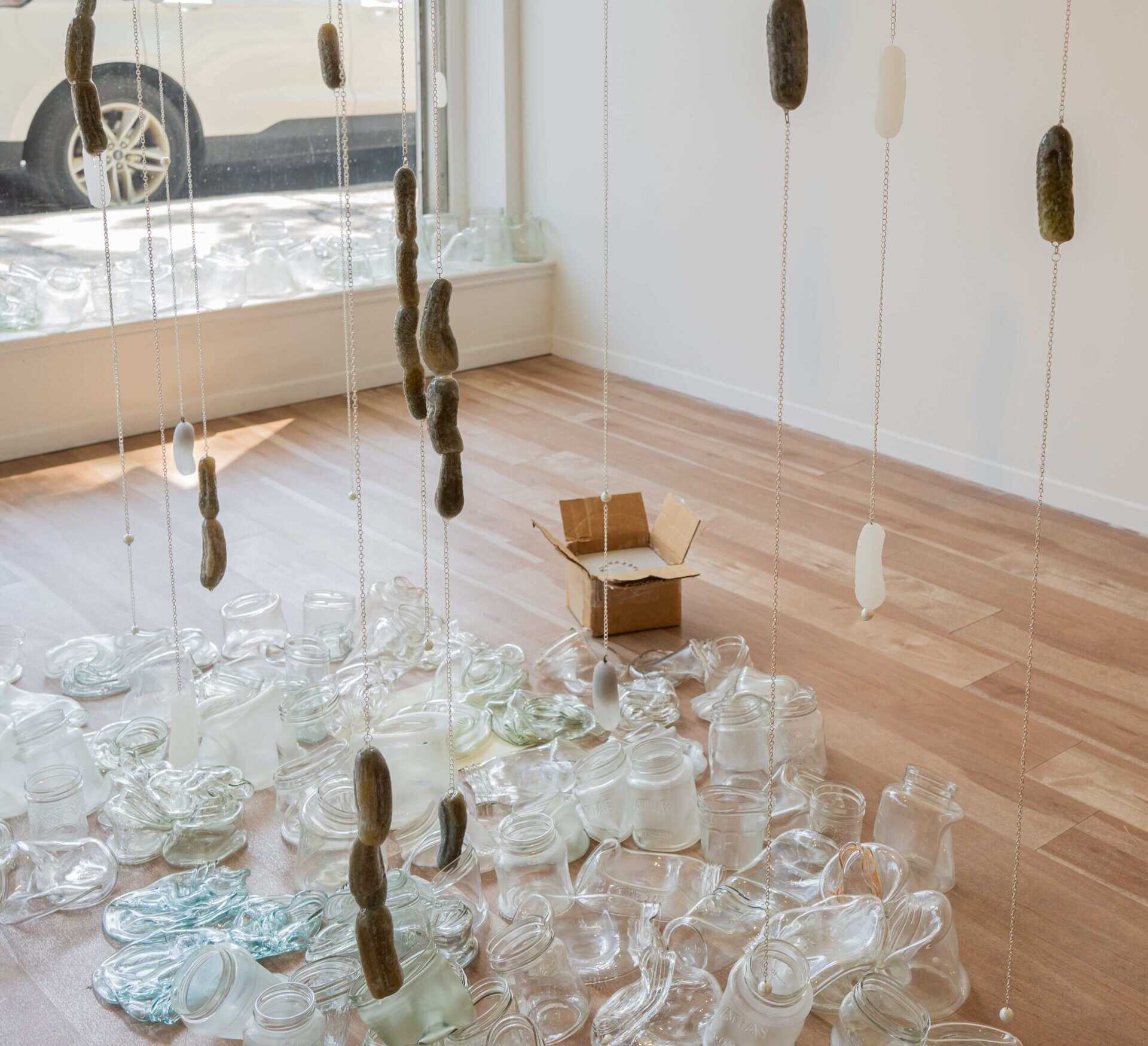 An art installation features glass pickles hanging from chains above scattered empty glass jars and lids on a wooden floor, with a small open cardboard box among the jars near a sunlit window.
