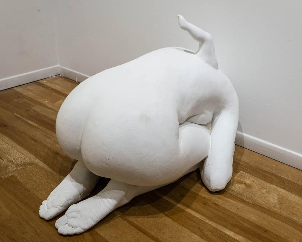 A white sculpture resembling a person positioned crouched on all fours in the corner of a gallery room with wooden floors and white walls.