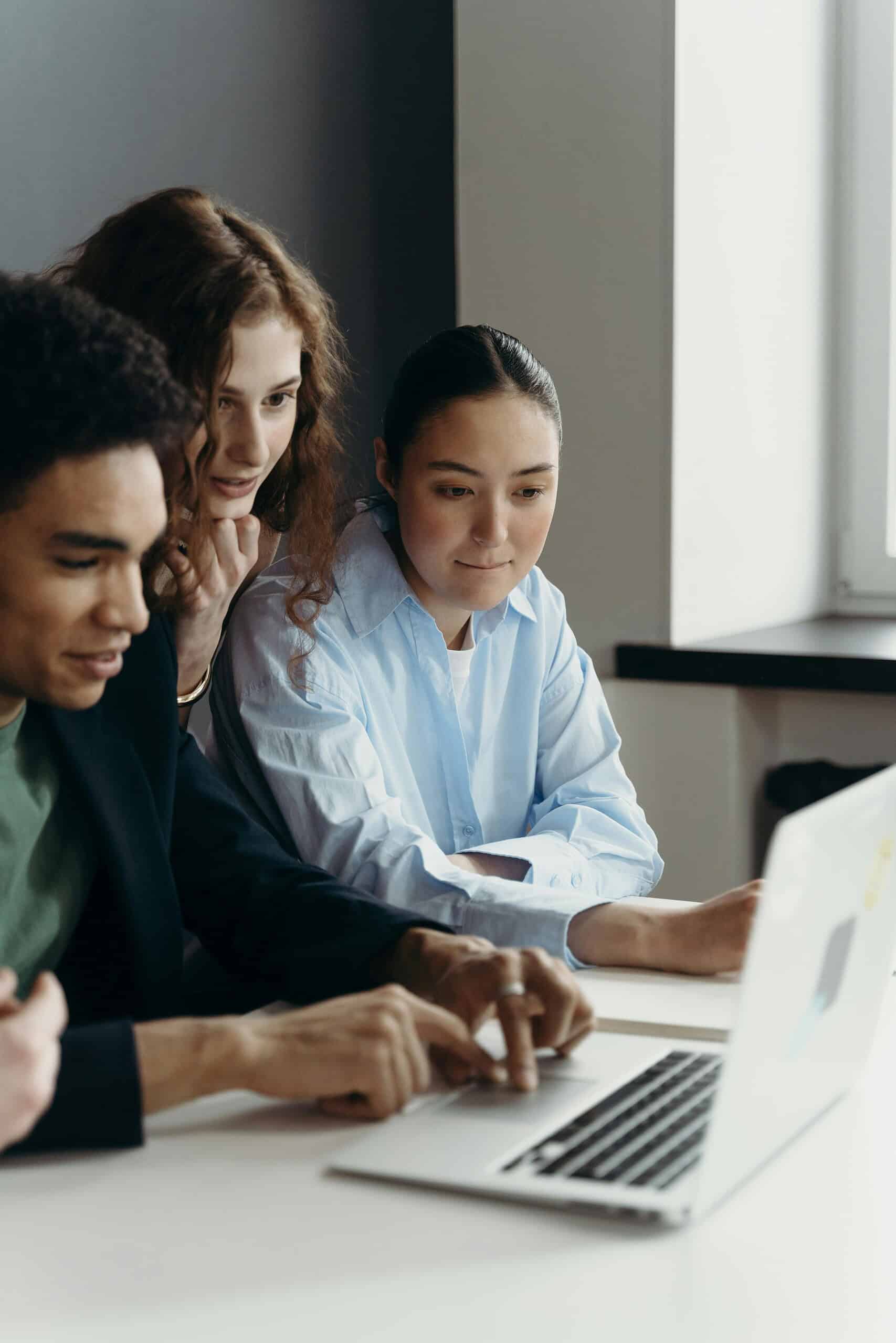 Three young people sit together at a table, focused on a laptop screen. One types while the others look on intently, highlighting collaboration and teamwork.