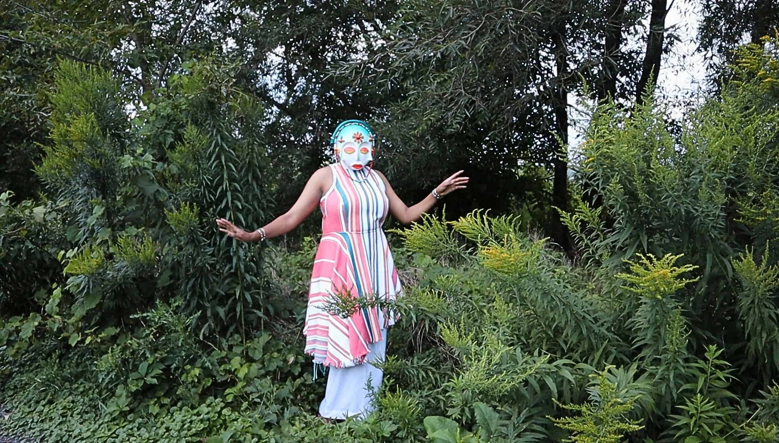 Person wearing a colourful striped dress and a decorative mask stands with arms raised among dense green foliage outdoors
