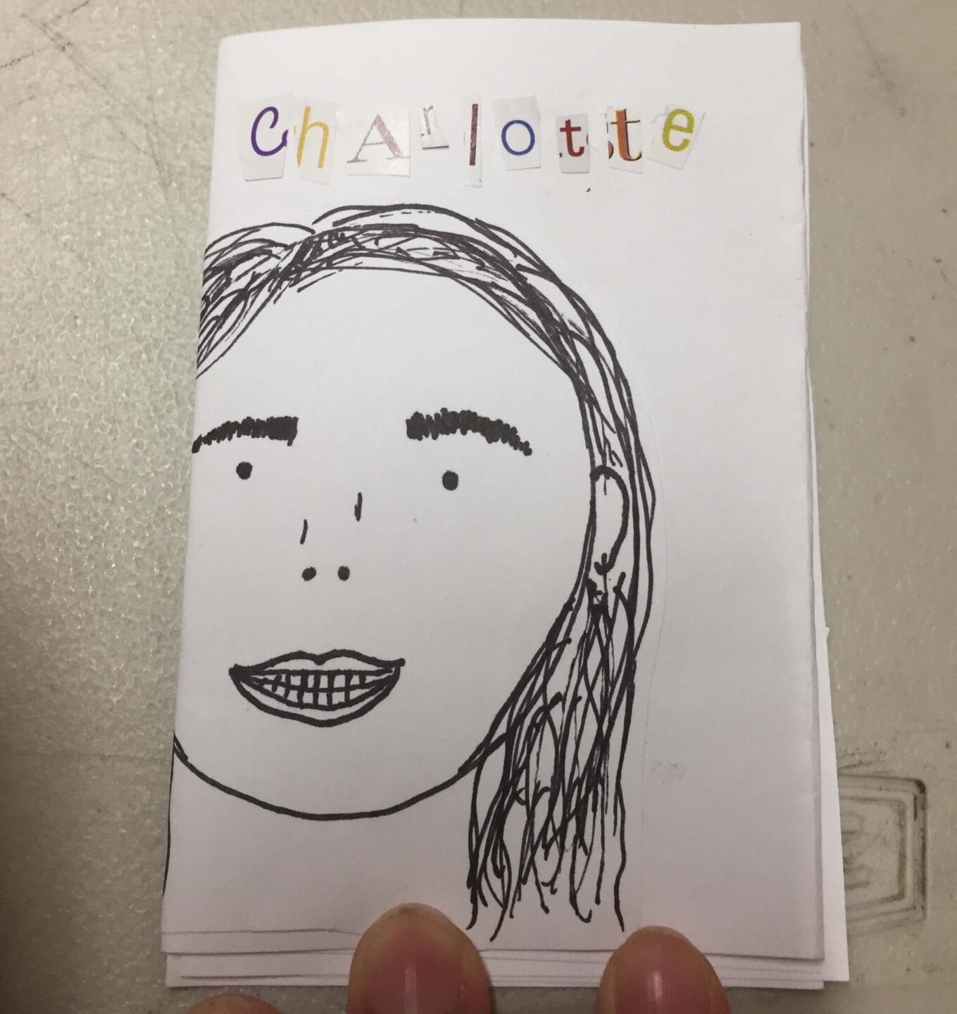 A handmade art booklet features a black marker drawing of a womans face on the cover. Above, Charlotte is spelled out in colorful cut-out magazine letters. Two fingers hold the booklet at the bottom, ready to enrich any collection.