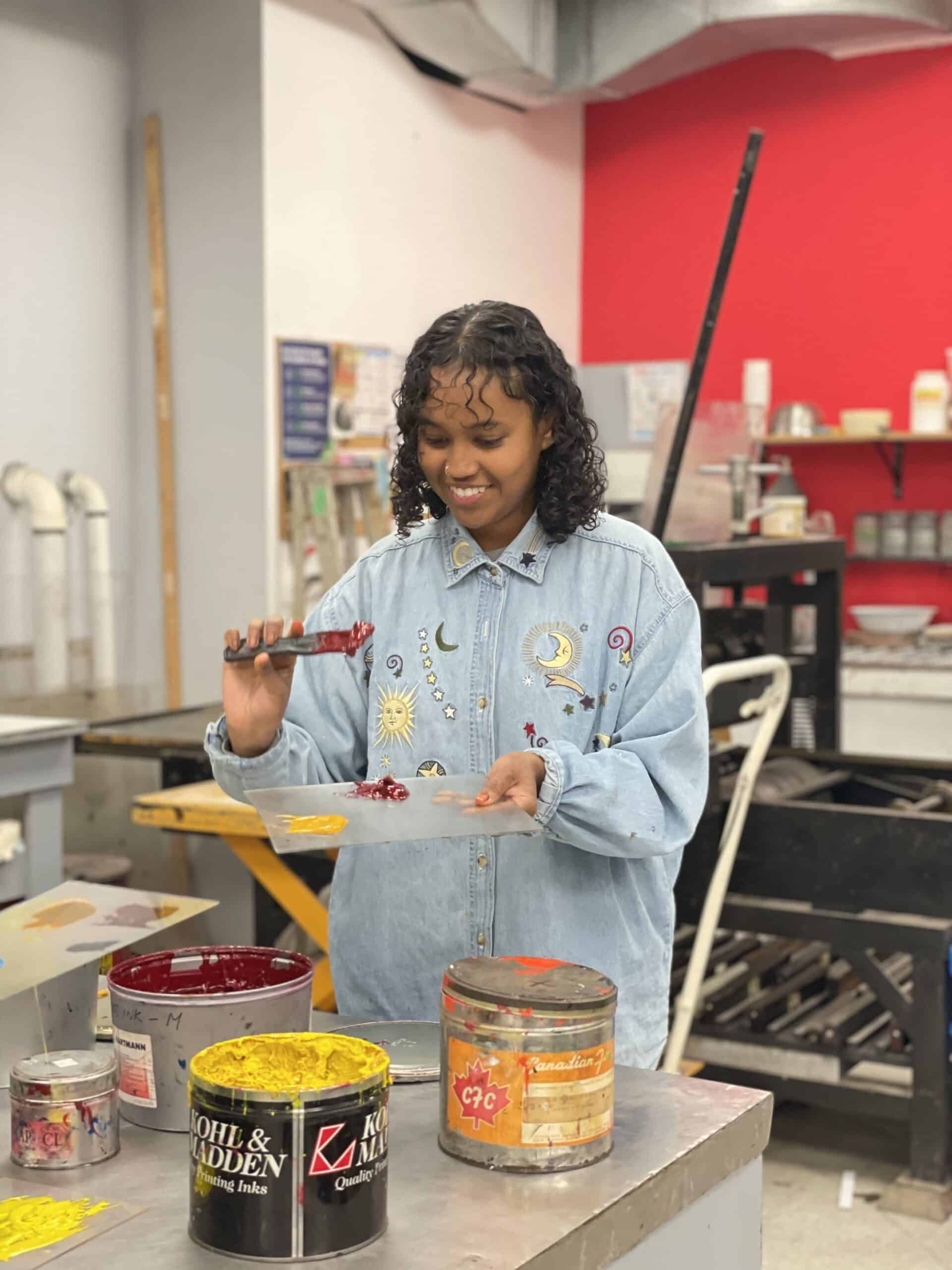 An artist wearing a decorated denim shirt smiles while spreading red ink on a palette in Centre[3]'s Traditional Printmaking Studio. Cans of yellow and red ink are on the table in front of them, and art supplies and printing presses are visible in the background.