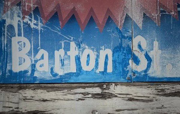A weathered blue Barton Street sign with distressed white lettering and a red zigzag pattern at the top.