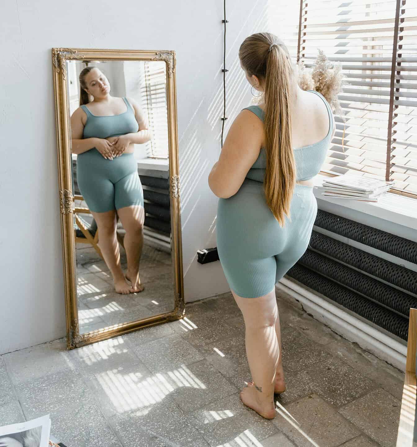 A young person looks at themselves critically in a full-body mirror.