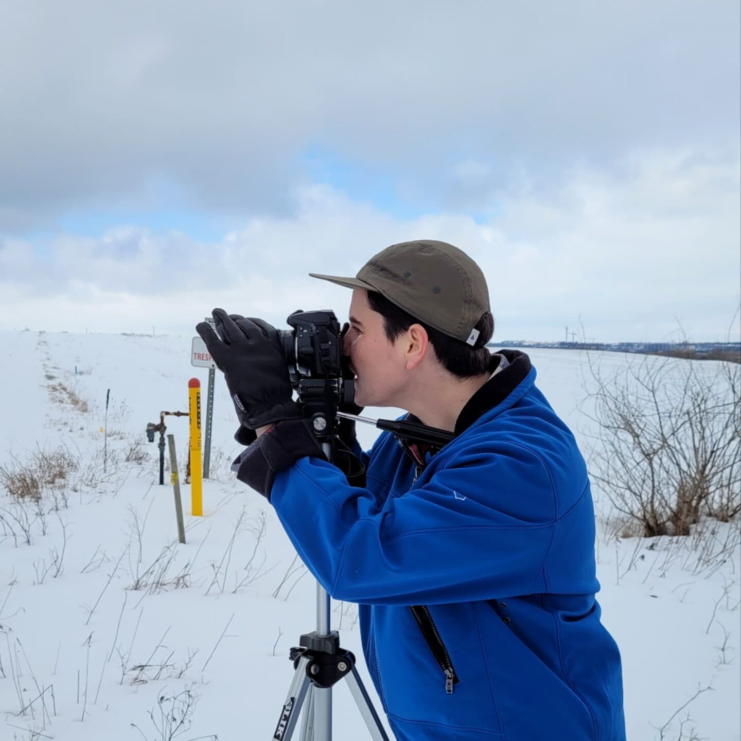 A Centre[3] resident artist in a blue jacket and grey cap uses a camera on a tripod to capture a photo in a snowy, open field beneath cloudy skies.