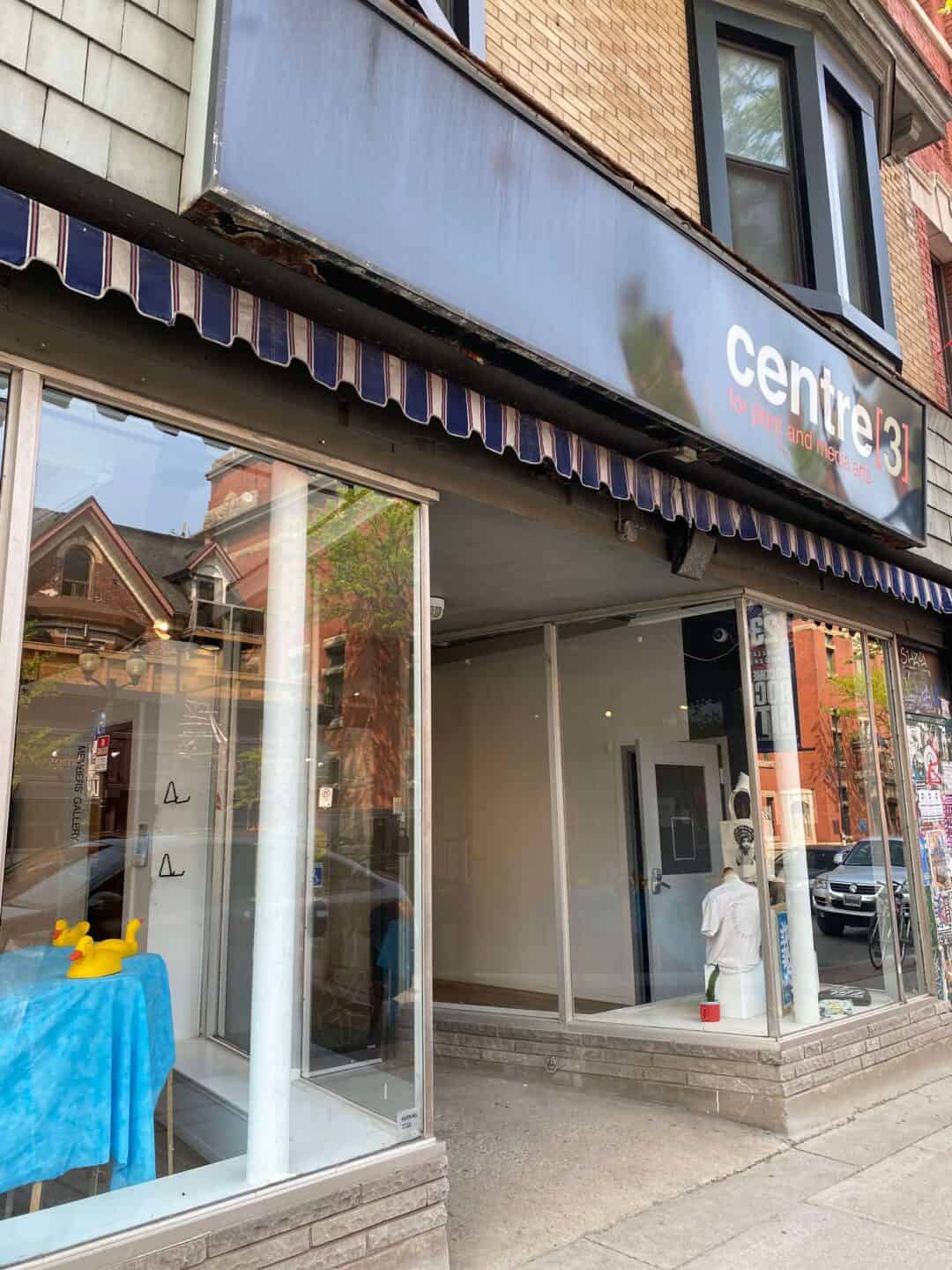 An outside street view of Centre[3] with a blue and white awning and sidewalk in view. A blue table with yellow plastic ducks and a white sculpture are inside the gallery windows. Brick buildings and cars line the street outside.