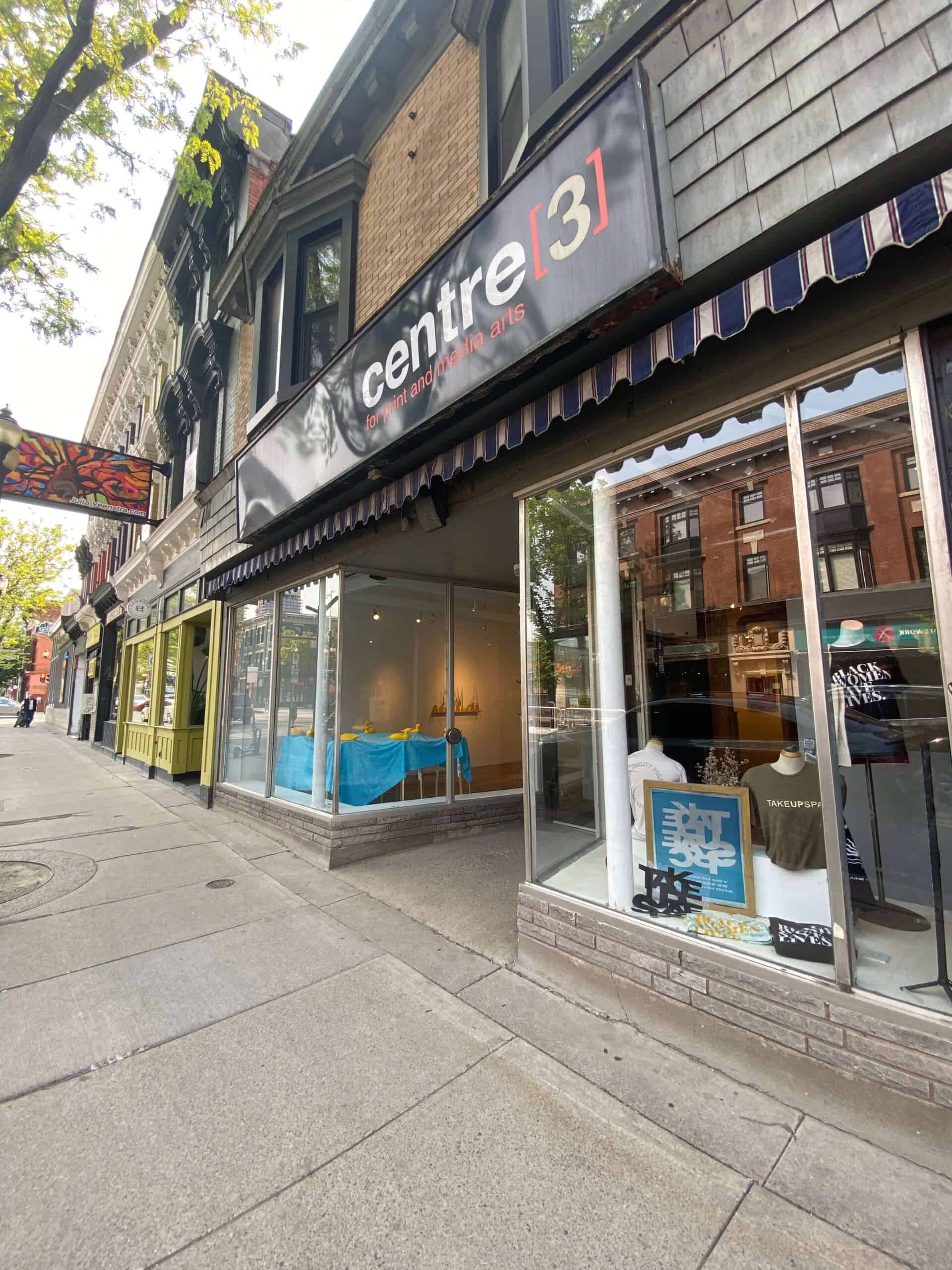 A wide sidewalk view of the Centre[4]Arts gallery front with large windows displaying artwork, brick buildings, and trees lining the street.