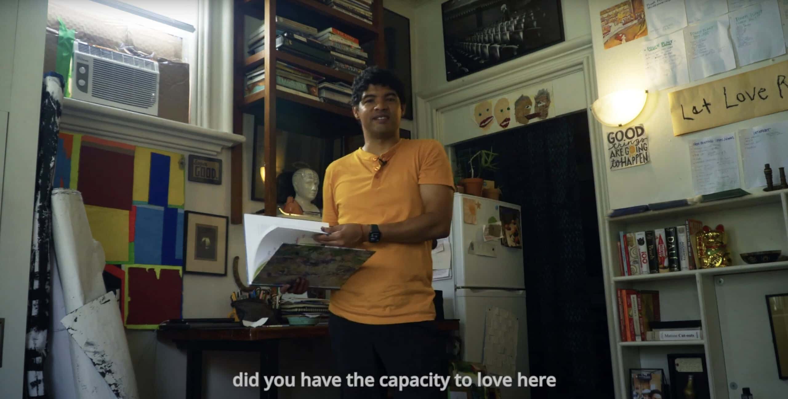 A person in an orange shirt stands in a cozy, art-filled room holding papers. Books, art supplies, and colourful notes fill the space.