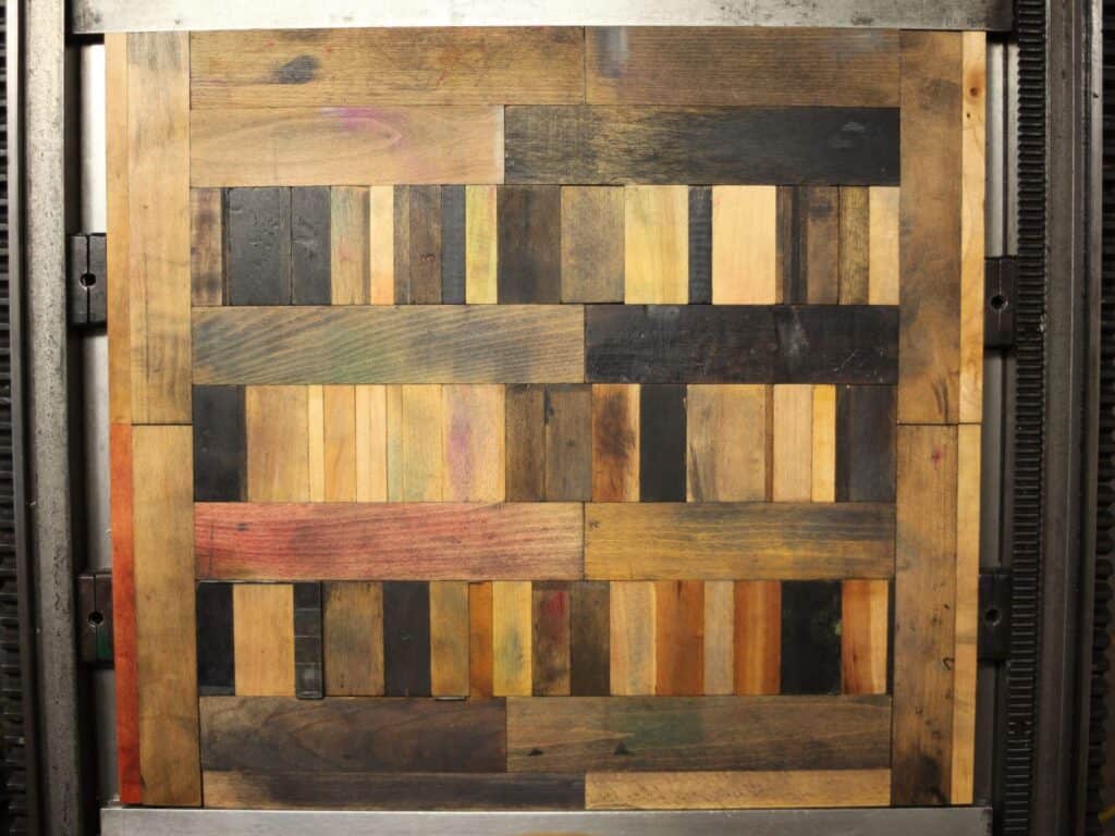 A square wooden panel made of rectangular cuts in various natural shades, arranged in a geometric, patchwork pattern with vertical and horizontal lock ups, all framed by a metal border.