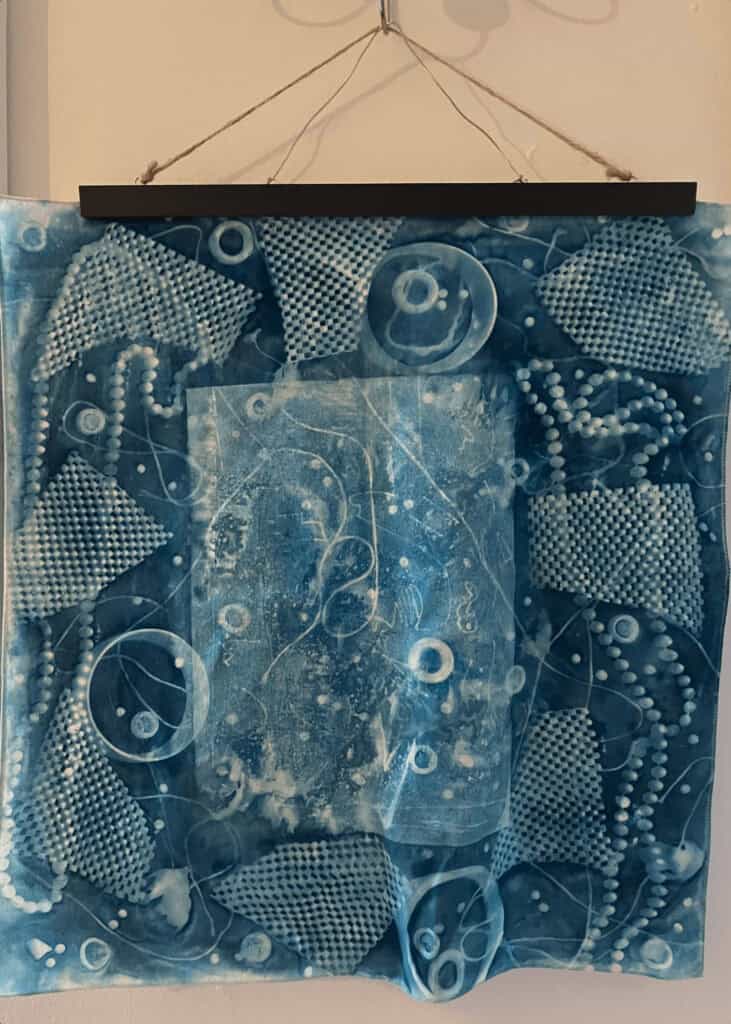 A blue and white abstract textile art piece with circular, dotted, and textured patterns is displayed hanging by a string and black bar against a plain wall. The layered design has an organic feel.