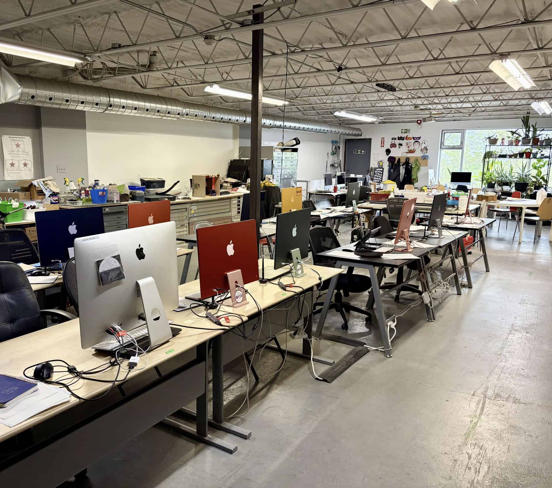 Rows of colourful iMac computers in Centre[3]'s Digital Lab. The tidy room, with scattered papers at some desks and large windows, is brightened by plants and natural light.
