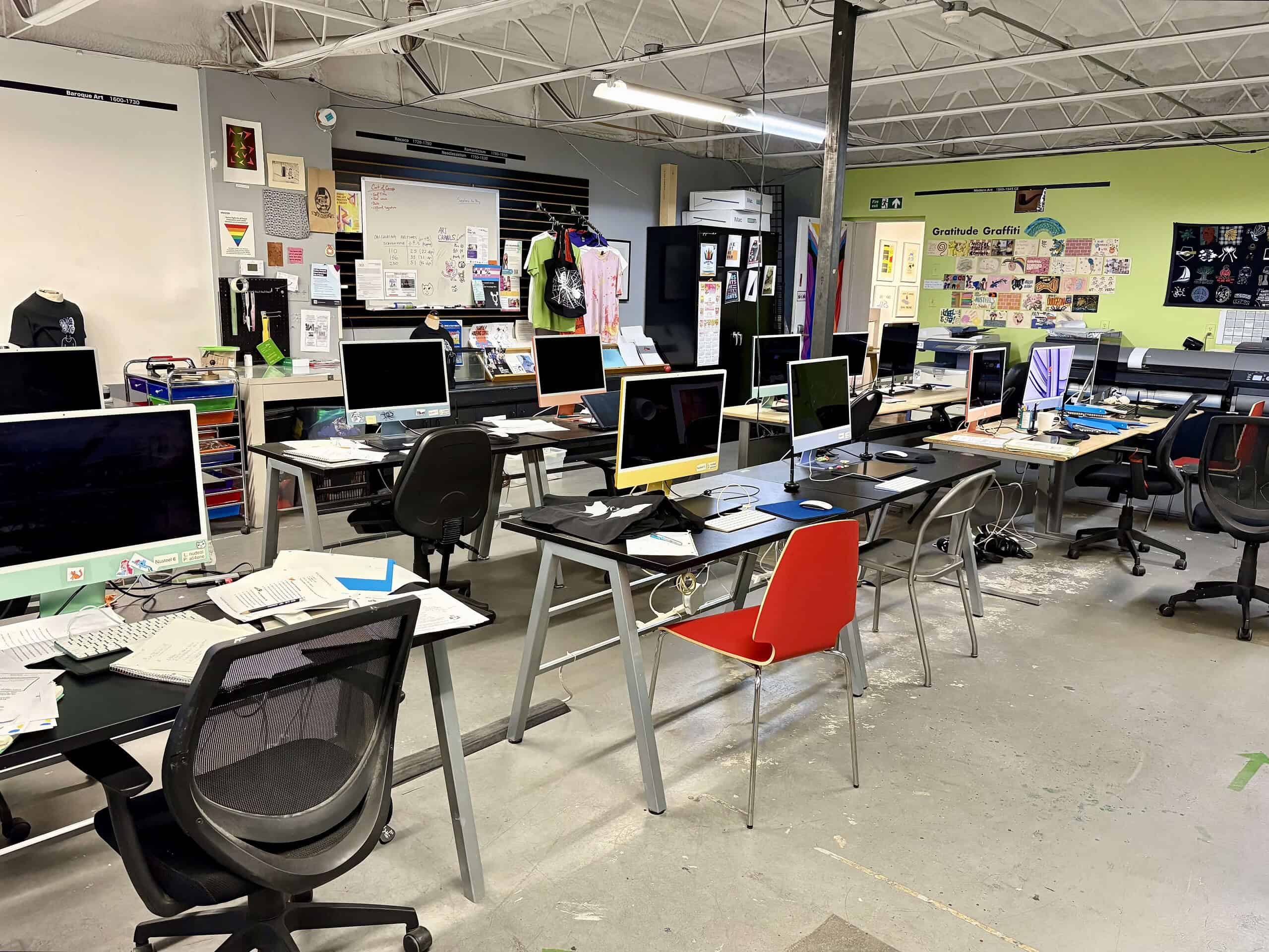 The Digital Computer Lab classroom at Centre[3] filled with desks, computers, scattered papers, and creative posters. The space appears active and slightly cluttered, with various supplies and equipment visible throughout.