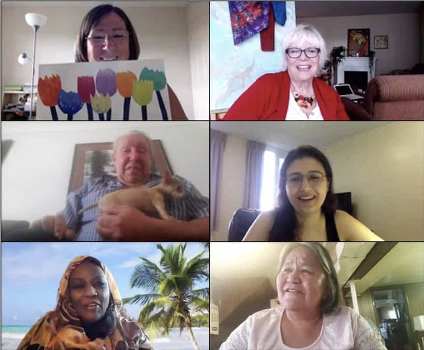 A screenshot from a video call with six adults, each in their own window. One holds up a colourful flower drawing; another holds a small dog on their lap, and one has a beach scene background. Everyone is smiling and looks content.