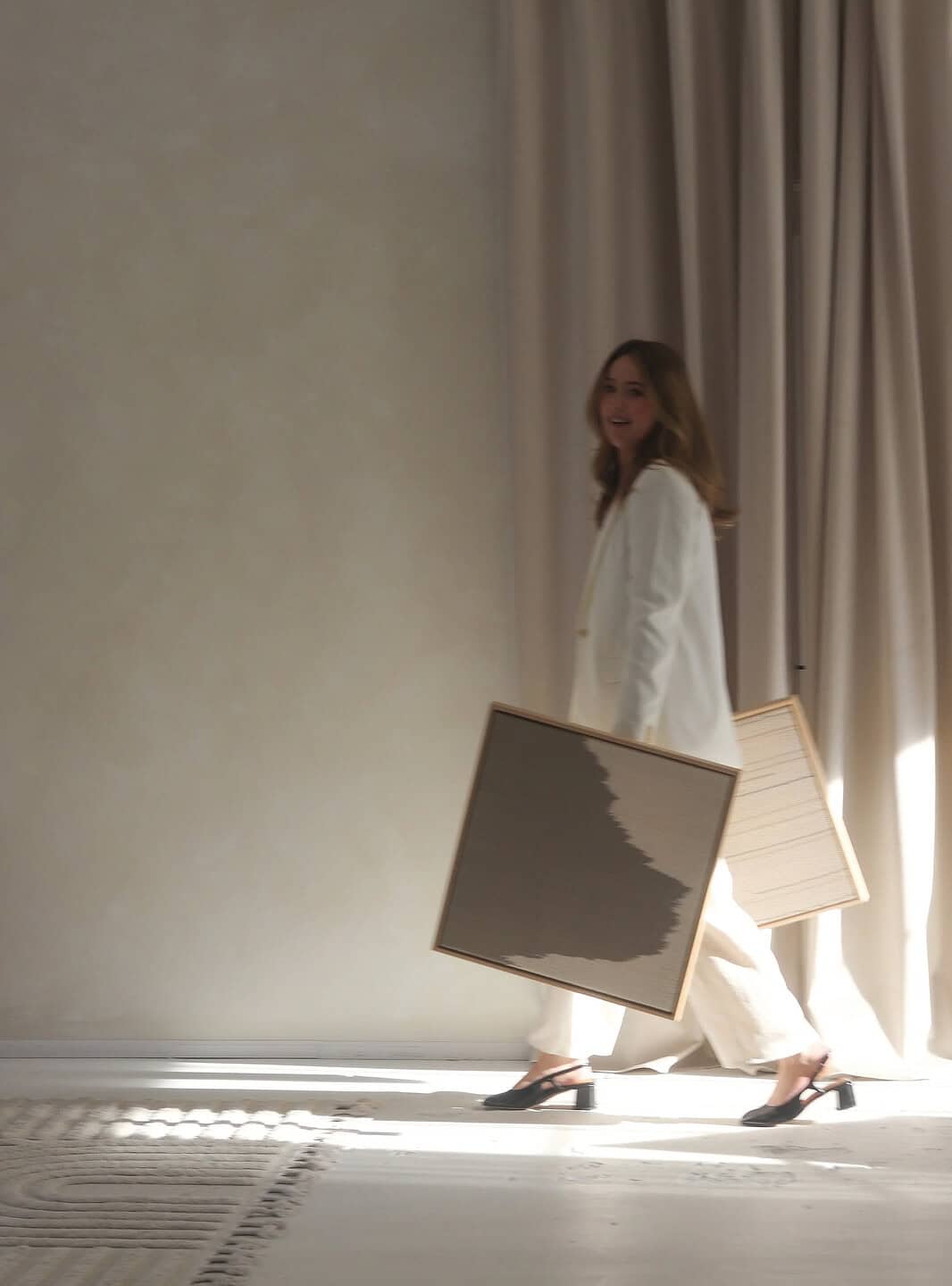 Emma Enright in a white outfit walks through a softly lit room carrying two framed artworks, with light streaming across the floor