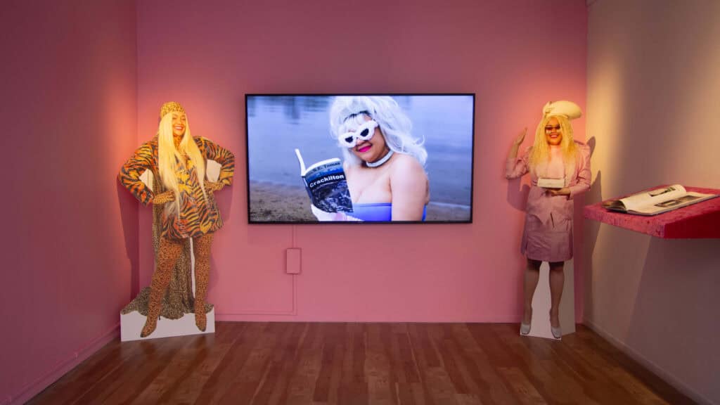 Two life-sized cardboard cutouts of people on either side of a screen showing the same person in a blonde wig, sunglasses, and blue top reading a book titled "Crackilon". A red velvet shelf with books is mounted on the right wall.