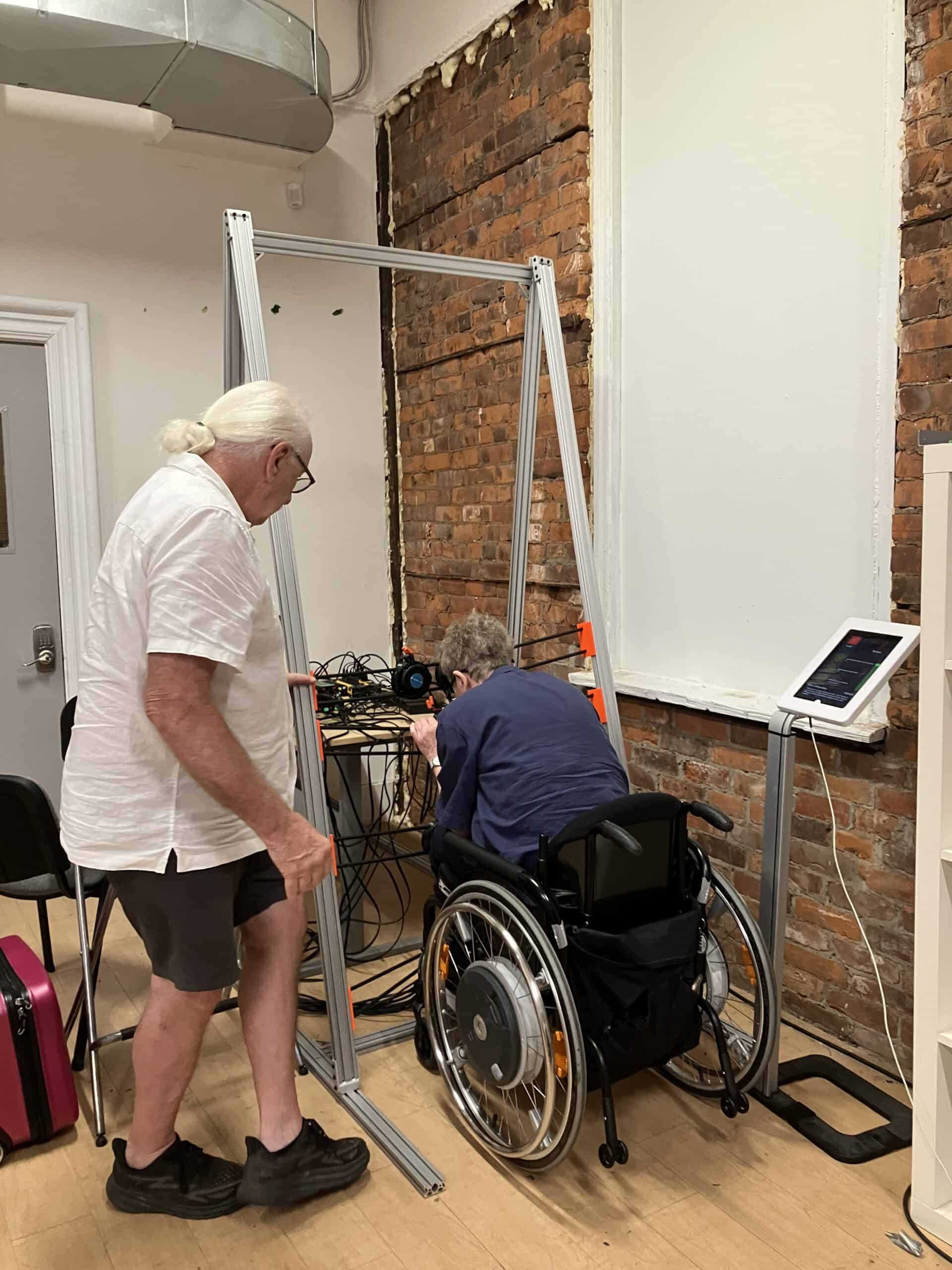 Two older adults set up electronic haptic equipment at the centre of a room with exposed brick walls; one is standing, while the other, in a wheelchair, adjusts cables inside a metal frame beside a nearby touchscreen device.
