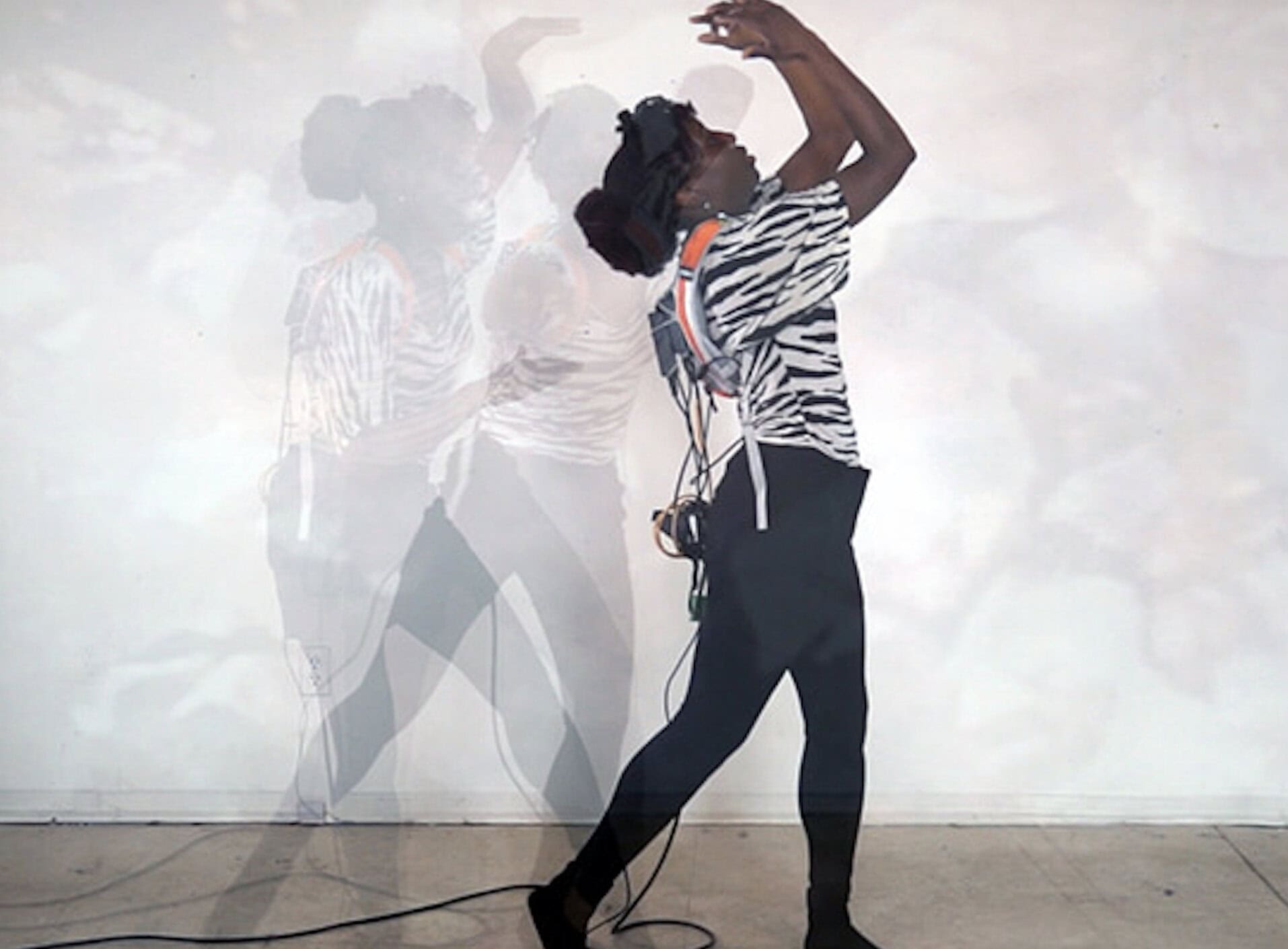 An individual wearing a zebra-striped shirt, black leggings, and a harness with wires attached performs a graceful, upward-reaching dance pose. Their movement is captured in layered, semi-transparent silhouettes, creating a sense of motion against a neutral wall and sidewalk.