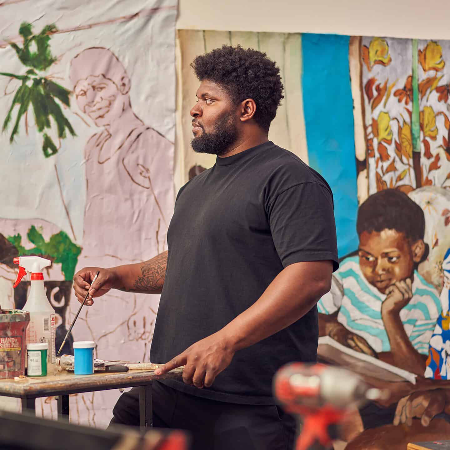 Artist Kareem-Anthony Ferreira stands at a work table holding a paintbrush, with large figurative paintings on fabric hanging behind them