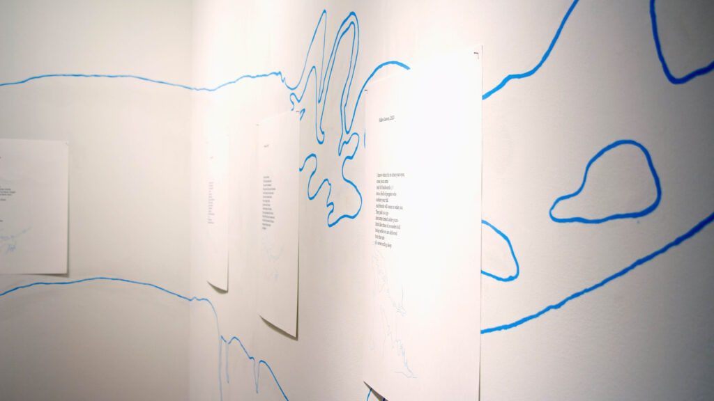 A gallery with three white papers featuring poems, displayed on a white wall. Thin blue lines are drawn across the wall, resembling abstract or map-like pathways.