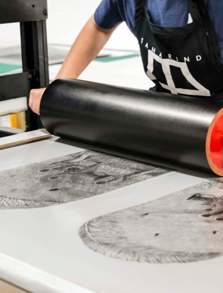 An artist uses a large roller to transfer a detailed black-and-white lithography print onto paper.